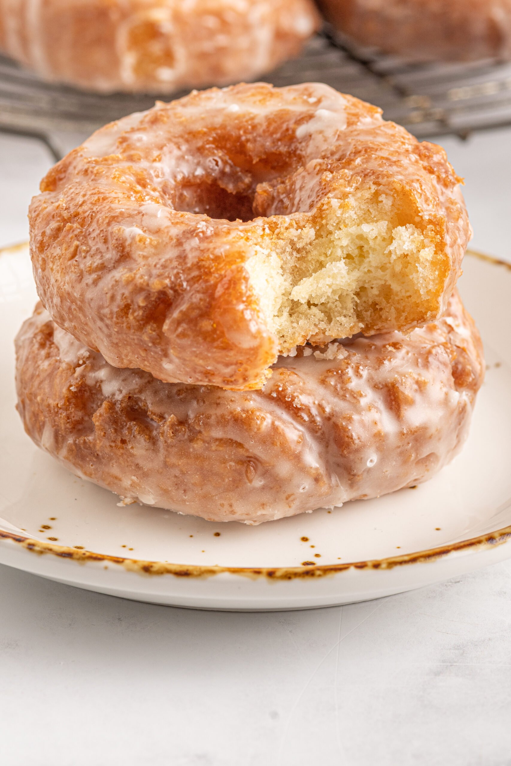 Two Old Fashioned Glazed Donuts stacked on a tan and brown plate with a bite removed from the top donut showing the fluffy inside. 