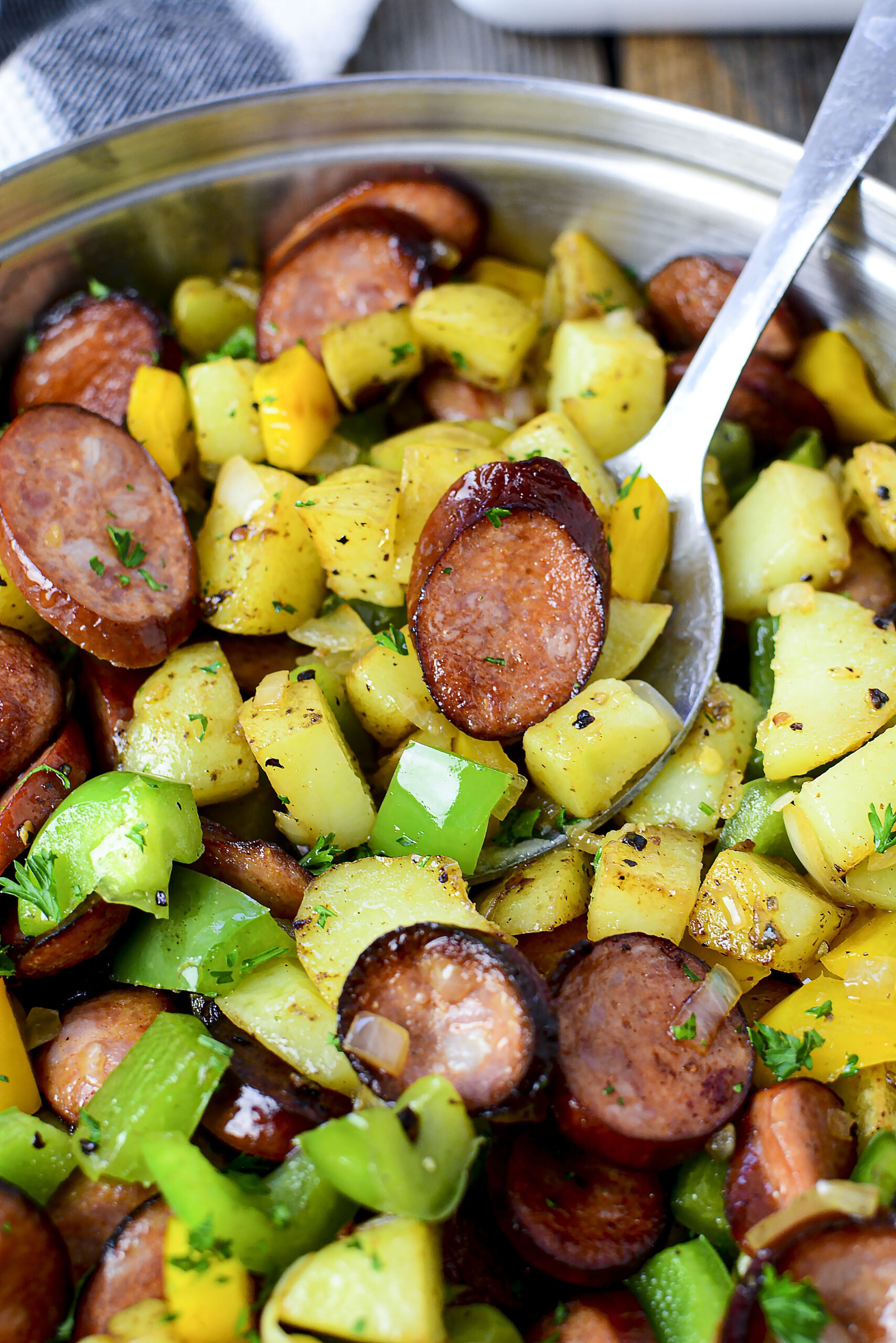 Overhead view of Sausage peppers and potatoes in a silver skillet with a spoon removing some for serving. 