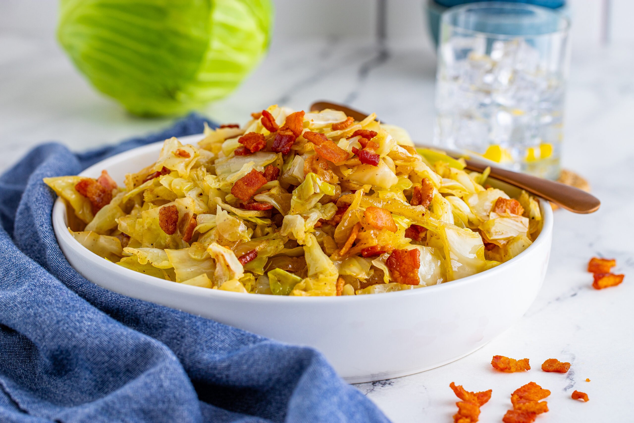 Horizontal image of bacon fried cabbage in a white bowl with a navy linen. 