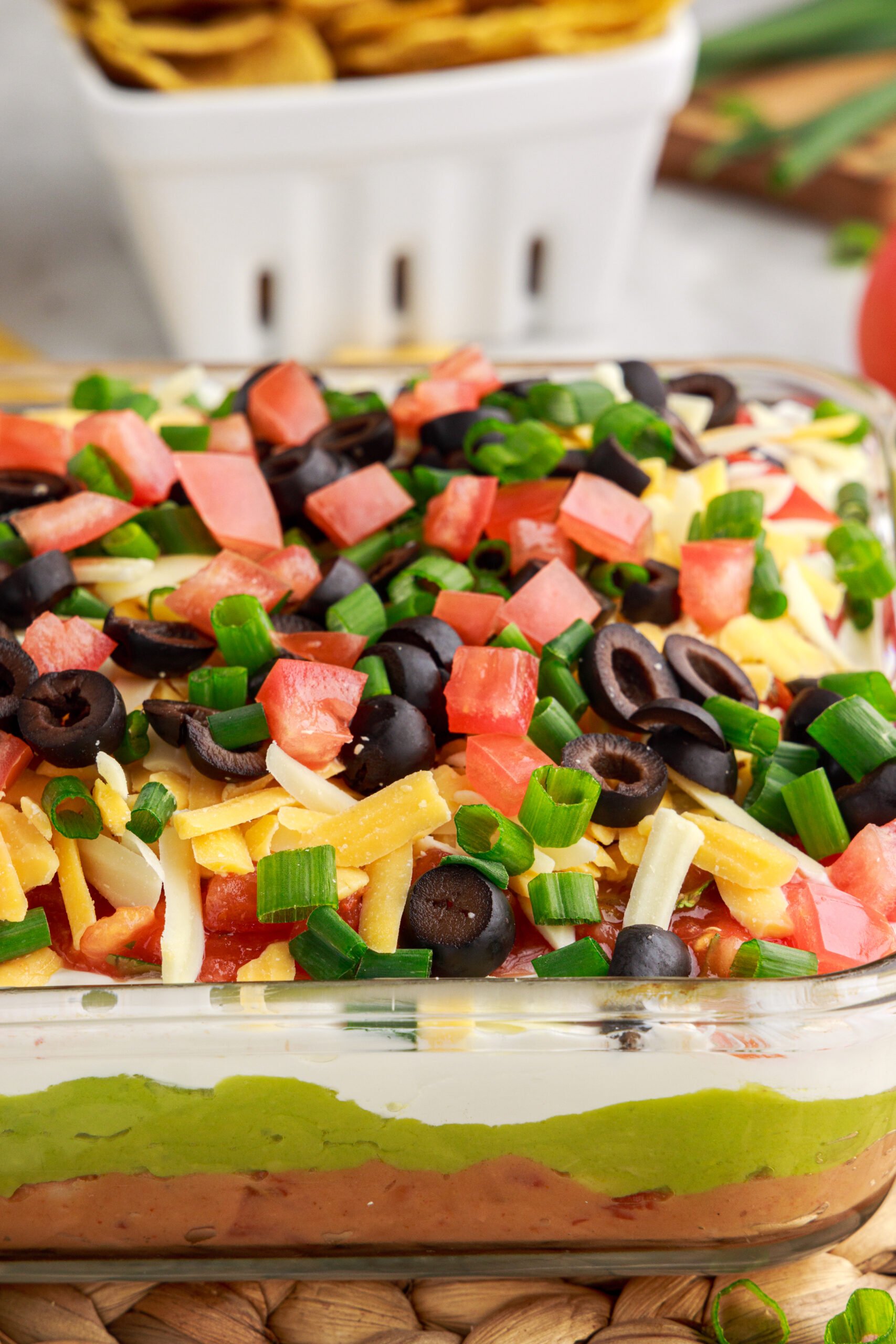 Close up view of 7-Layer Dip in a clear baking dish, topped with diced tomatoes , black olives, cheese and chives