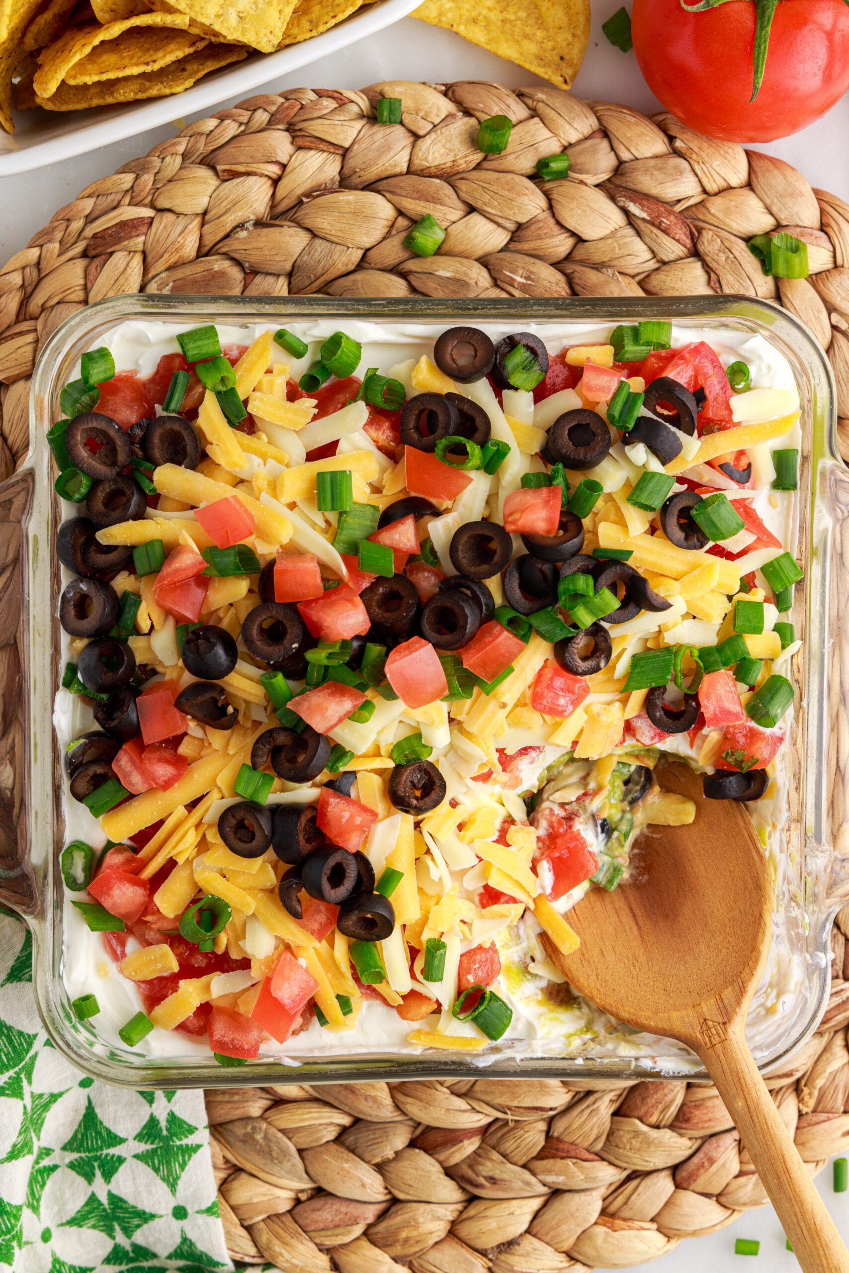 Overhead view of 7-Layer Dip in a clear baking dish with a serving removed by a wooden spoon.