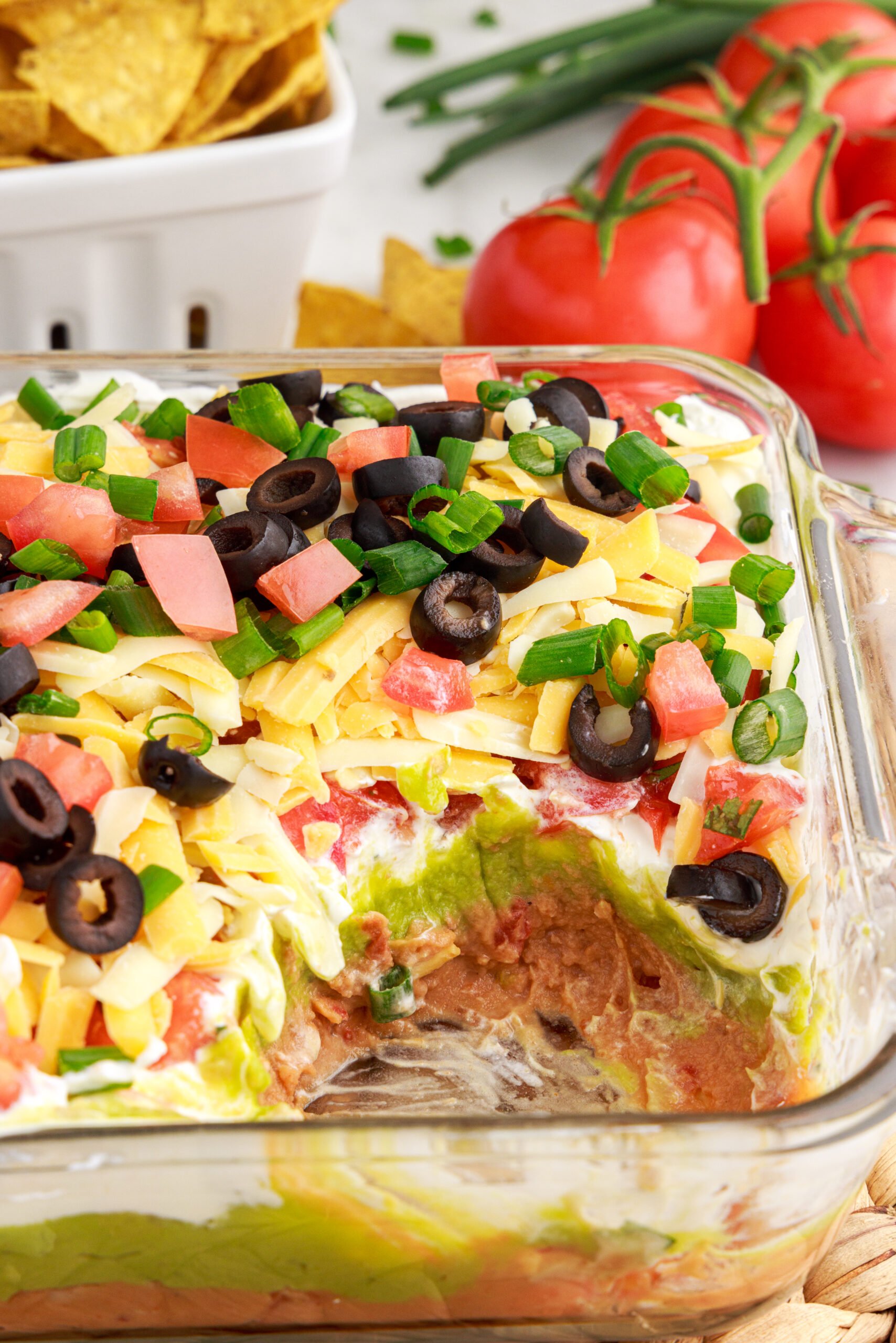 big scoop missing out of the 7-Layer Dip in a clear baking dish, topped with diced tomatoes , black olives, cheese and chives