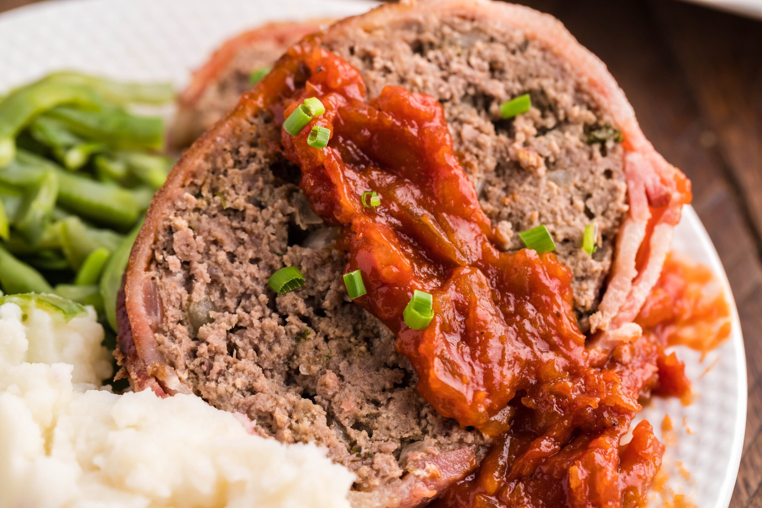 Close up view of a slice of Bacon Wrapped Meatloaf on a white dish served with mashed potatoes and green beans.