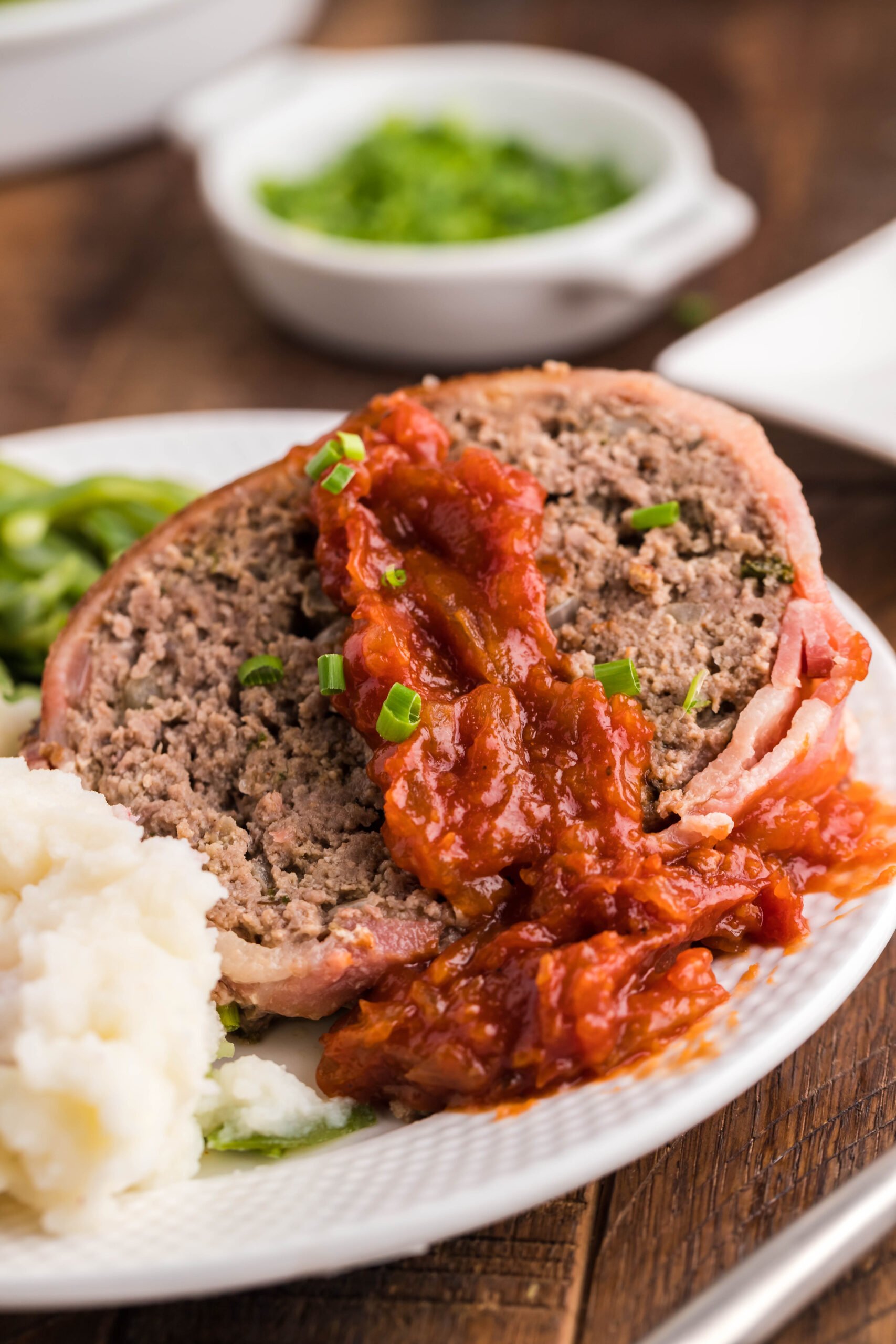 Close up view of a slice of Bacon Wrapped Meatloaf on a white dish served with mashed potatoes and green beans.