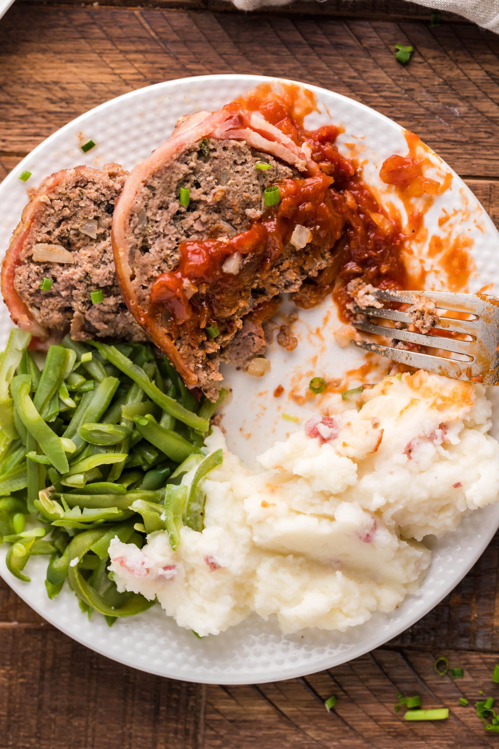 Overhead view of a slice of Bacon Wrapped Meatloaf, cut in half by a silver fork, on a white dish served with mashed potatoes and green beans.