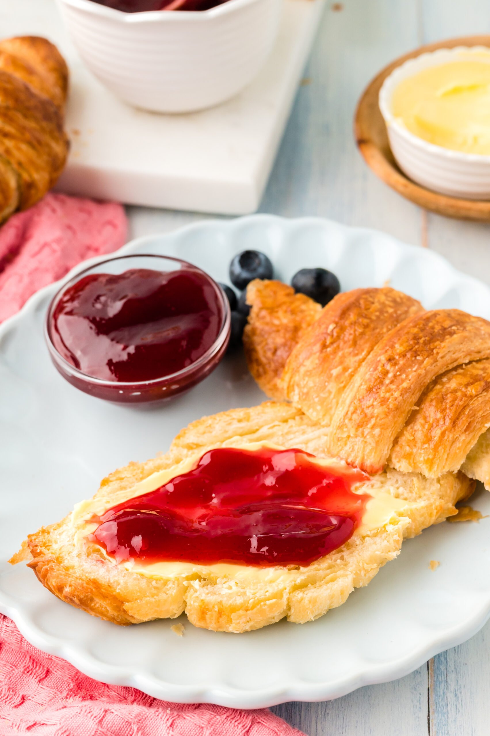 Front view of Homemade Croissants cut in half with jam spread on the bottom half, served on a white dish with jam and fresh blueberries.