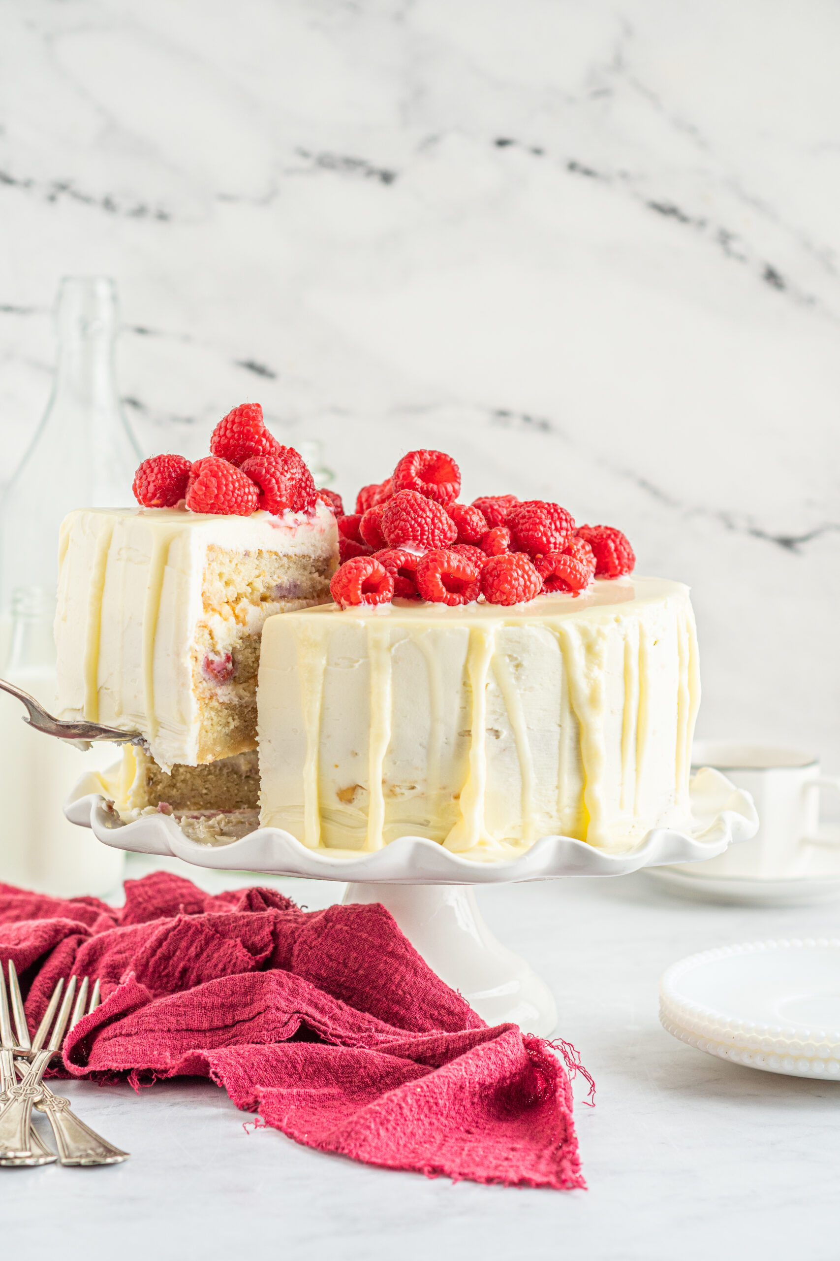 Front view of White Chocolate Cake with Raspberries with a slice being removed by a silver cake server.