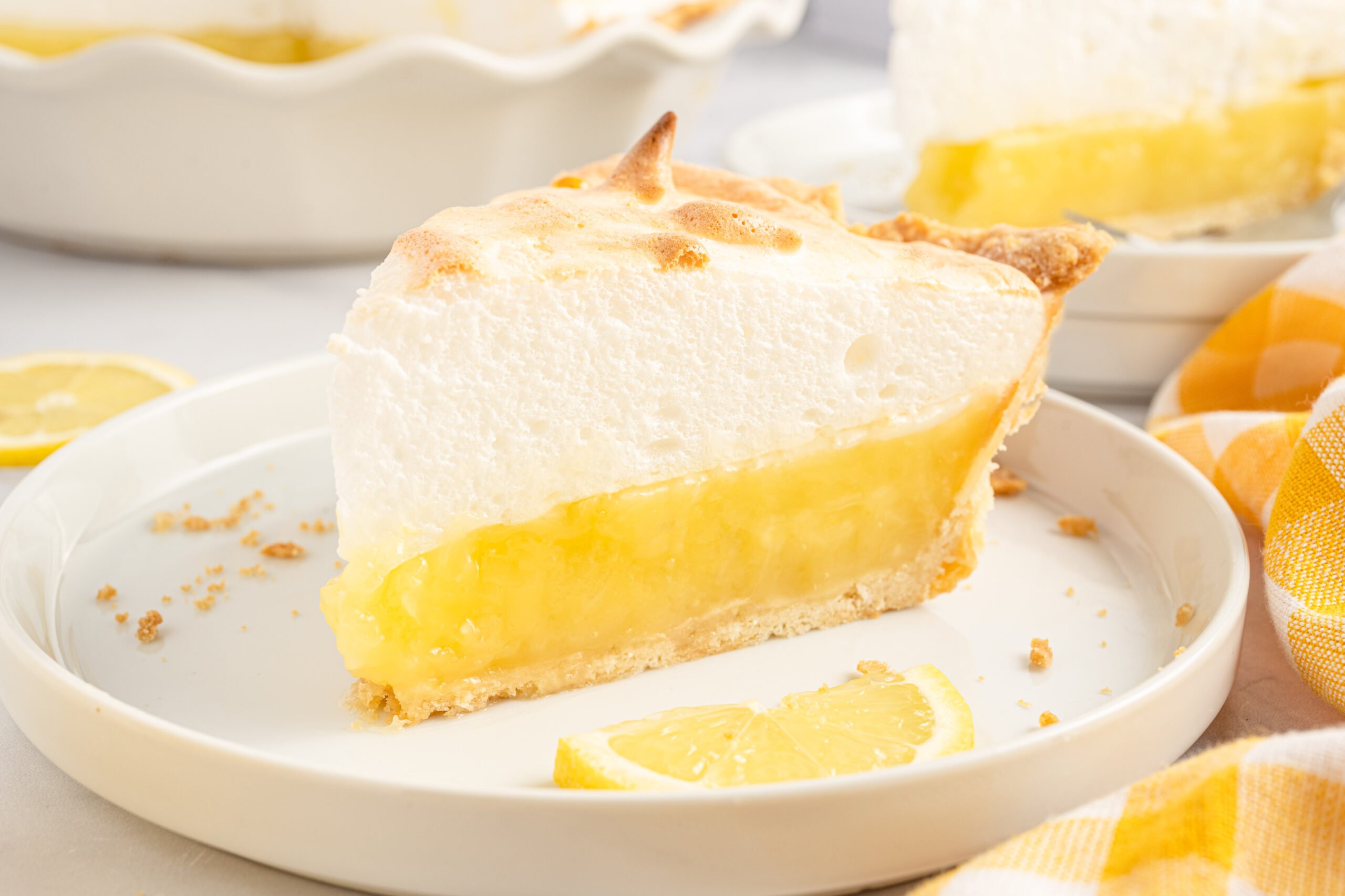 side view of a piece of the lemon meringue pie on a white plate with fresh sliced lemons. 
