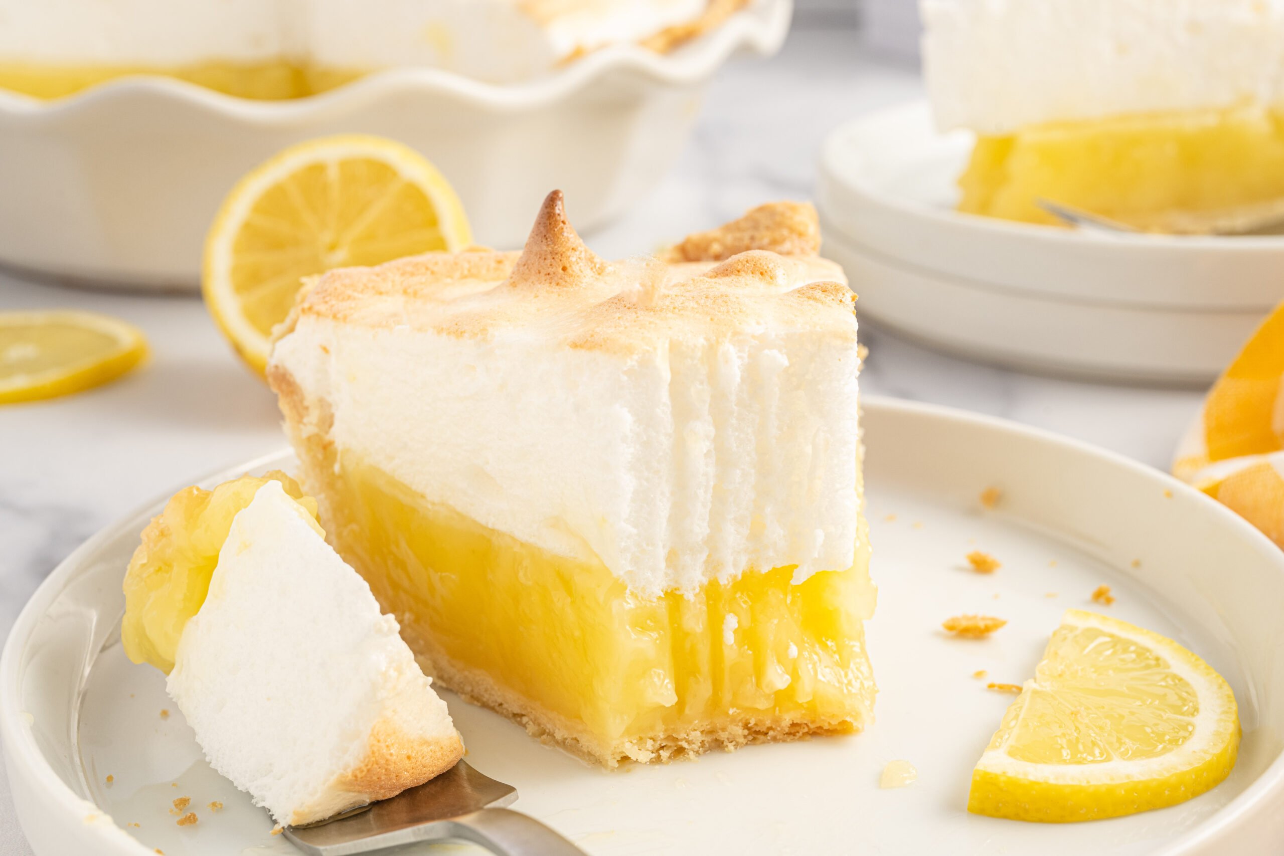 up close view of a piece of the lemon meringue pie with a bite missing on a white plate with fresh sliced lemons. 