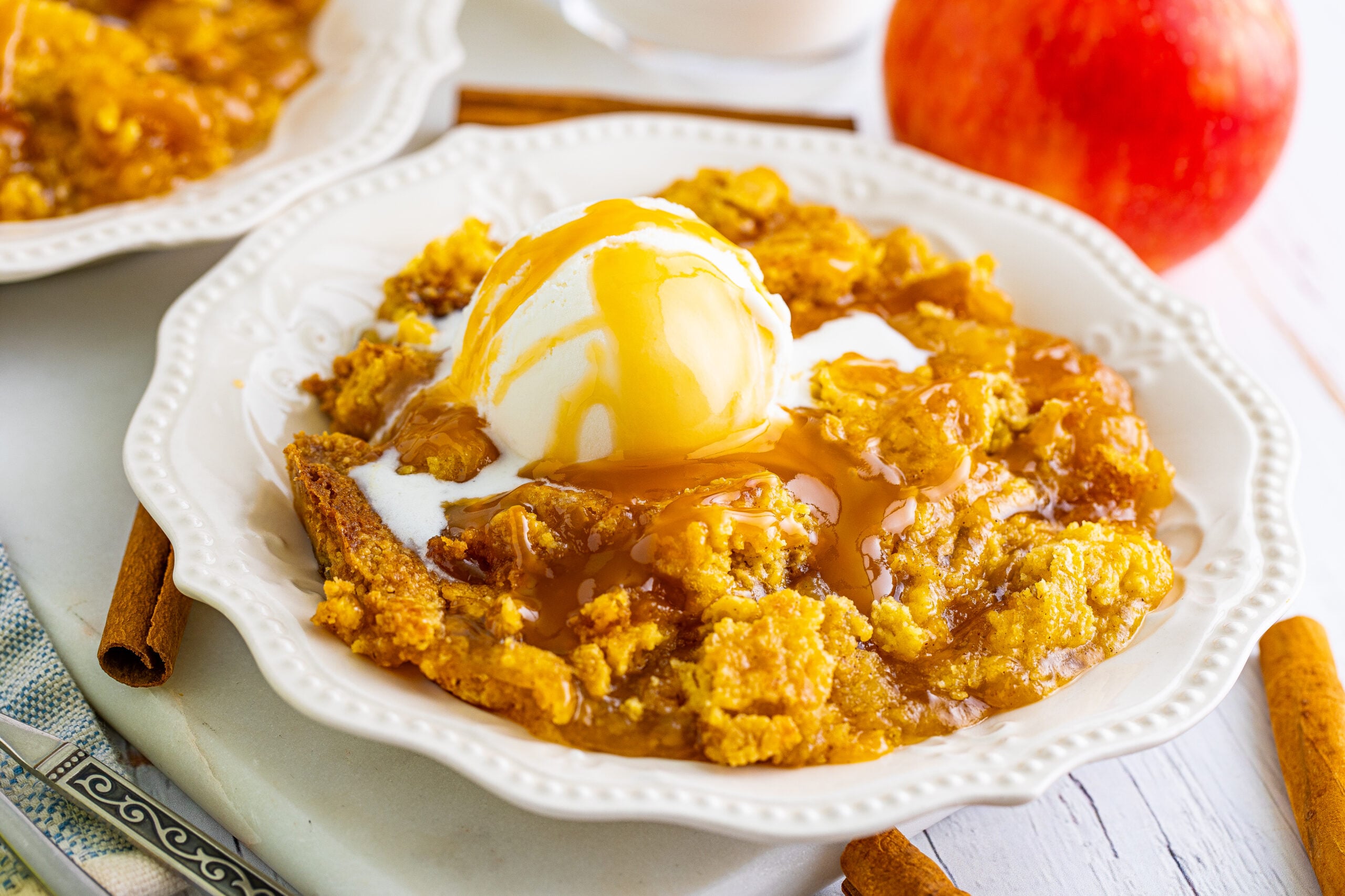 Close up shot of caramel apple dump cake on two white plates with cinnamon sticks and apples in the background.