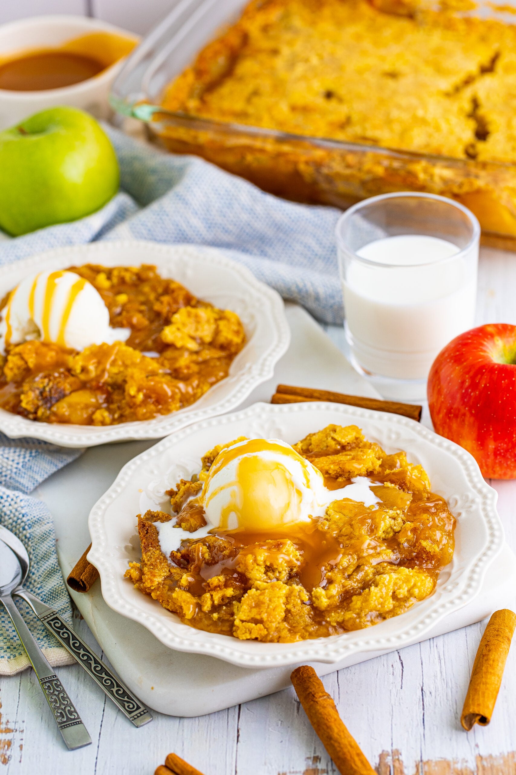 Close up shot of caramel apple dump cake on two white plates with cinnamon sticks and apples in the background.