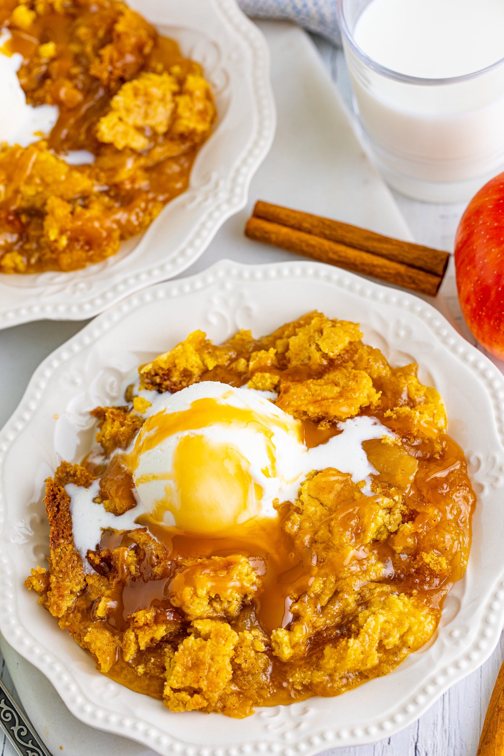Close up shot of caramel apple dump cake on two white plates with cinnamon sticks and apples in the background.