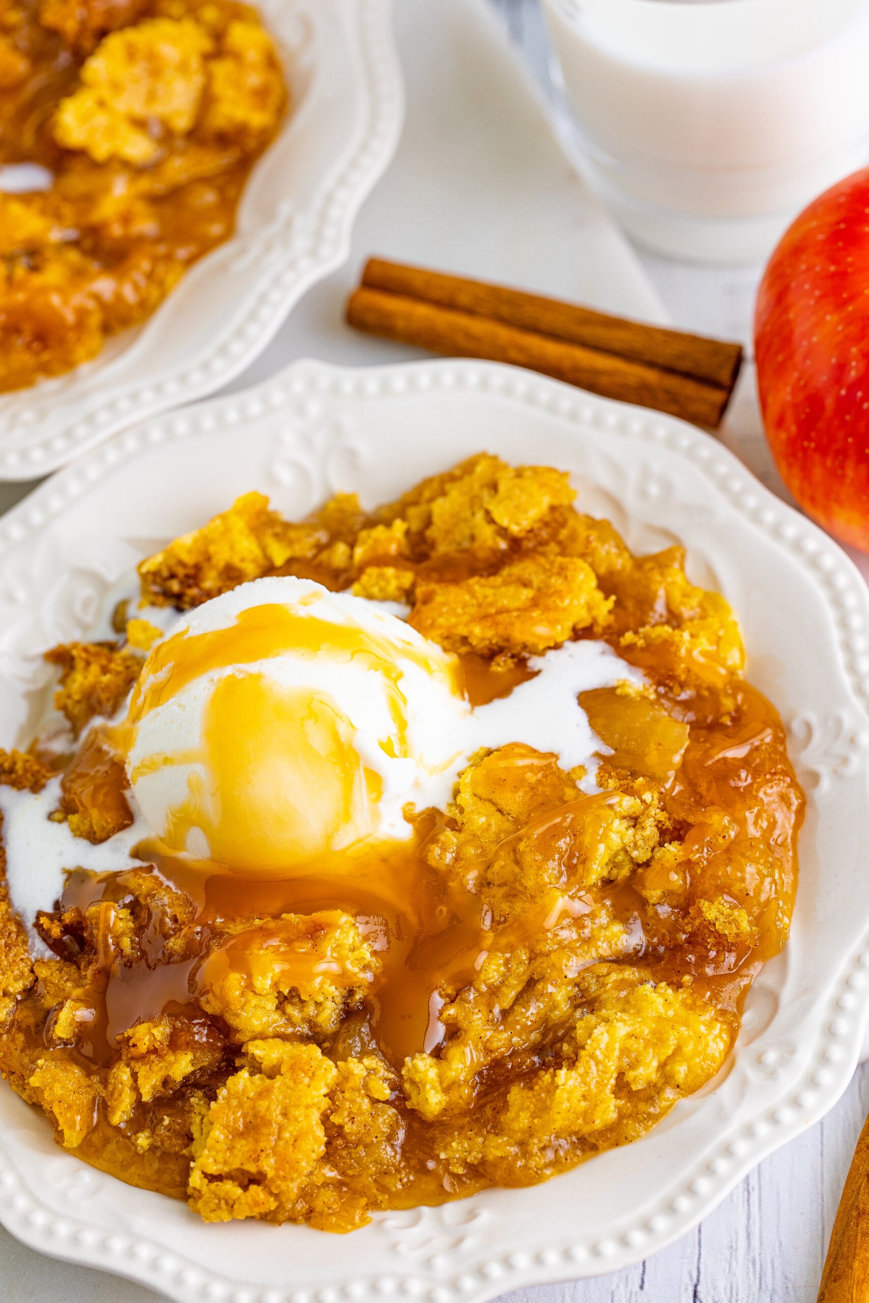 Close up shot of caramel apple dump cake on two white plates with cinnamon sticks and apples in the background.