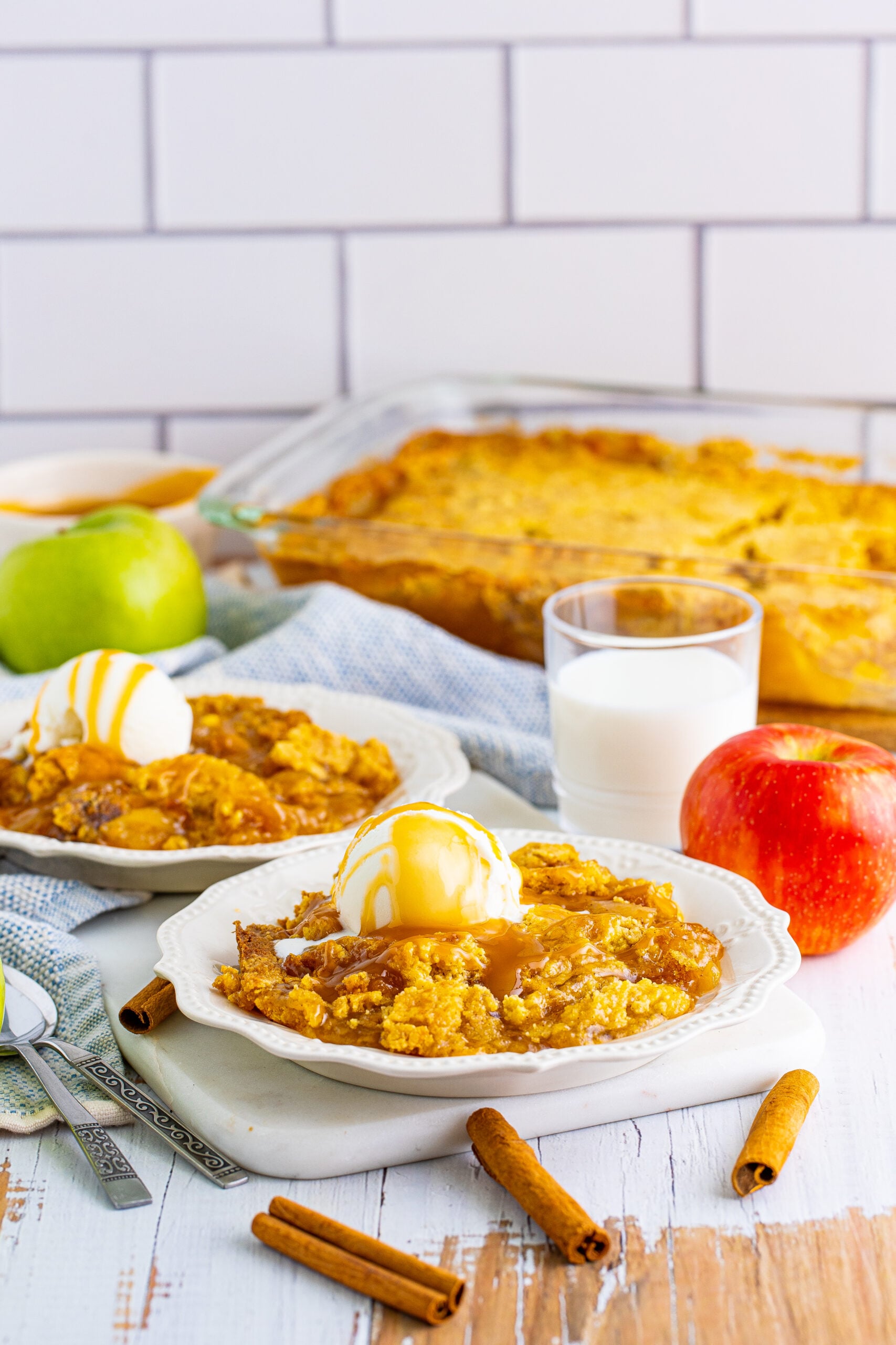Caramel apple dump cake on two white plates with cinnamon sticks and apples in the background.