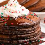 Hot Chocolate Pancakes stacked on a plate topped with syrp and whipped cream and holiday sprinkles