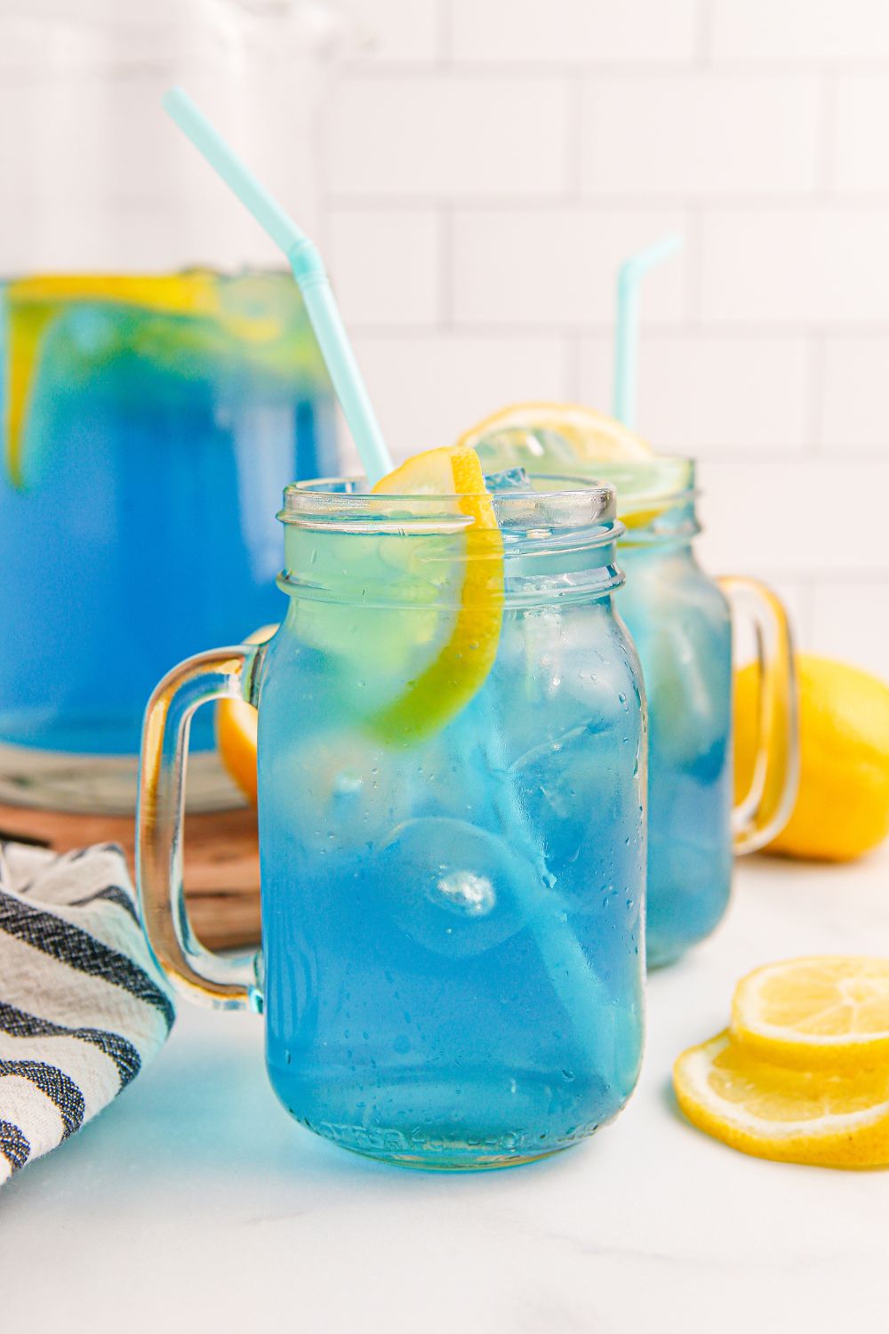 two glasses and a pitcher of Blue Raspberry Lemonade sitting on table with lemon slices around it on table 