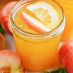 Peach Lemonade in a mason jar garnished with peach slice and orange slice. Fresh peaches around the glass