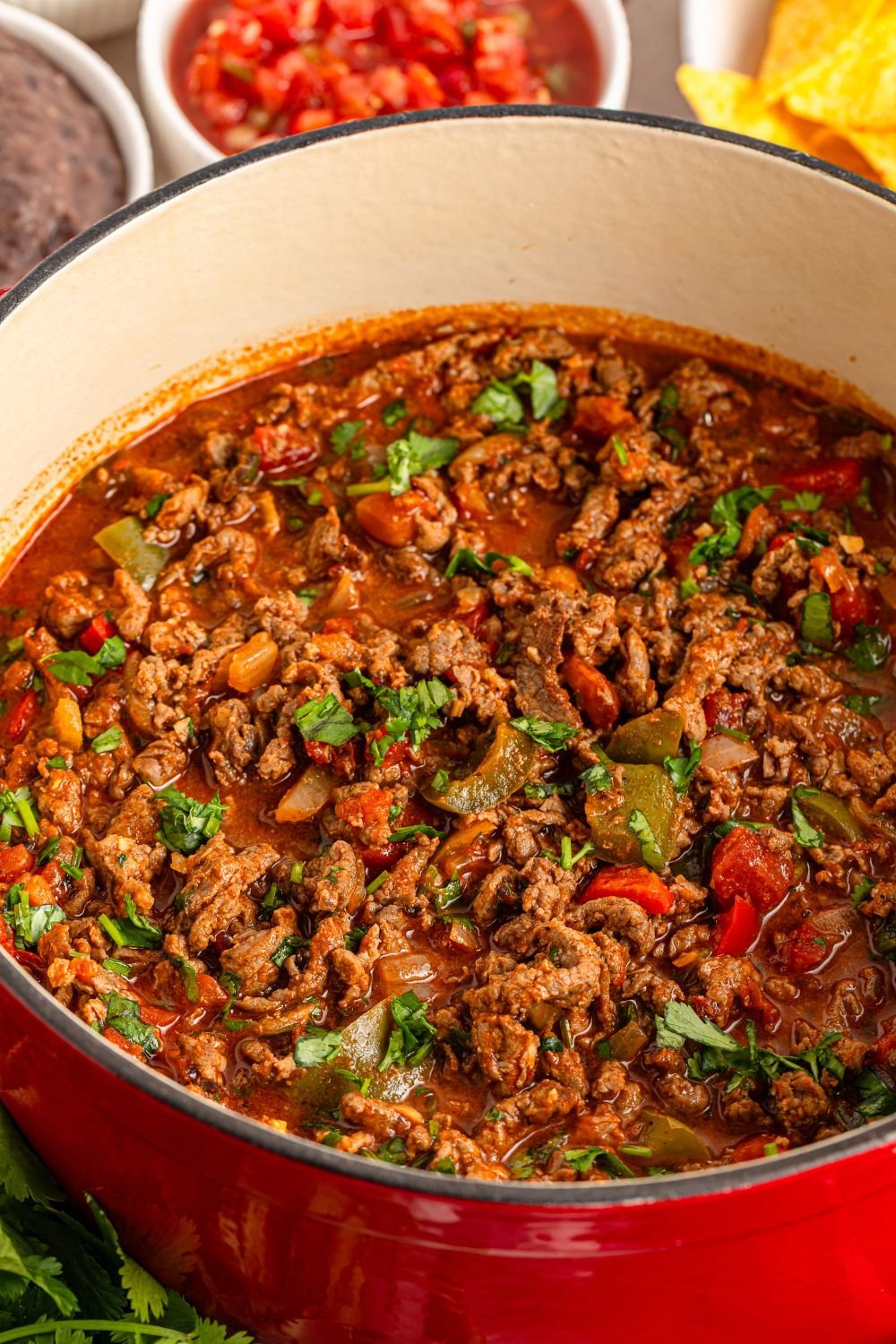 Carne Picada cooked in a Dutch oven with bowls of ingredients around the pan 