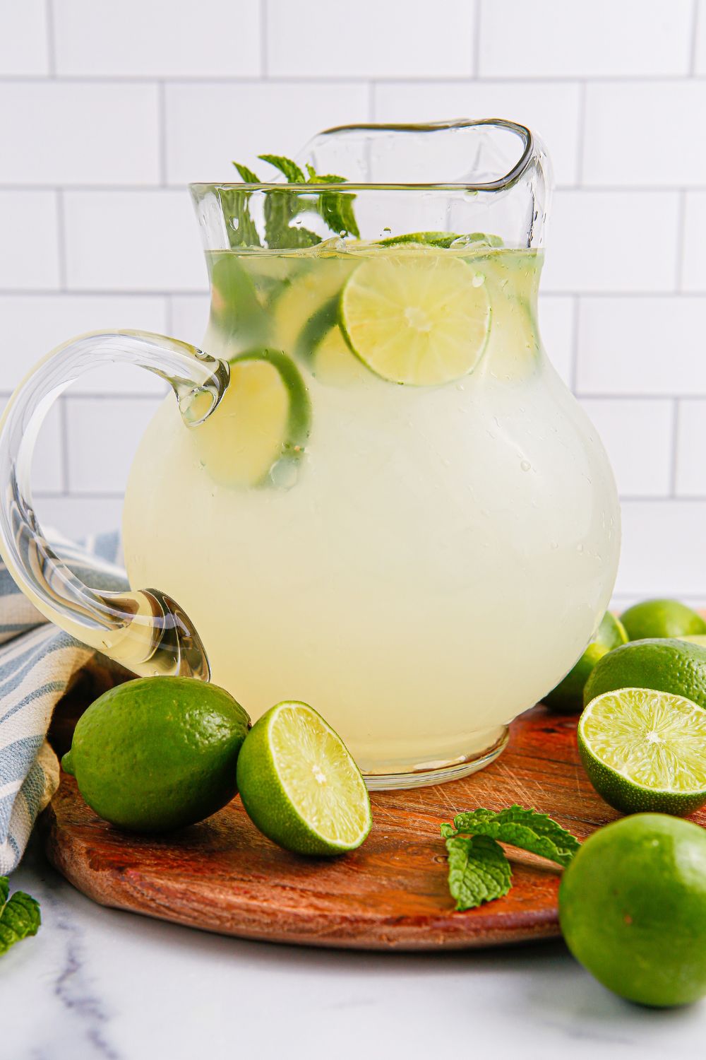 side view of limeade in a glass pitcher sitting on a wooden cutting board and limes placed around the pitcher
