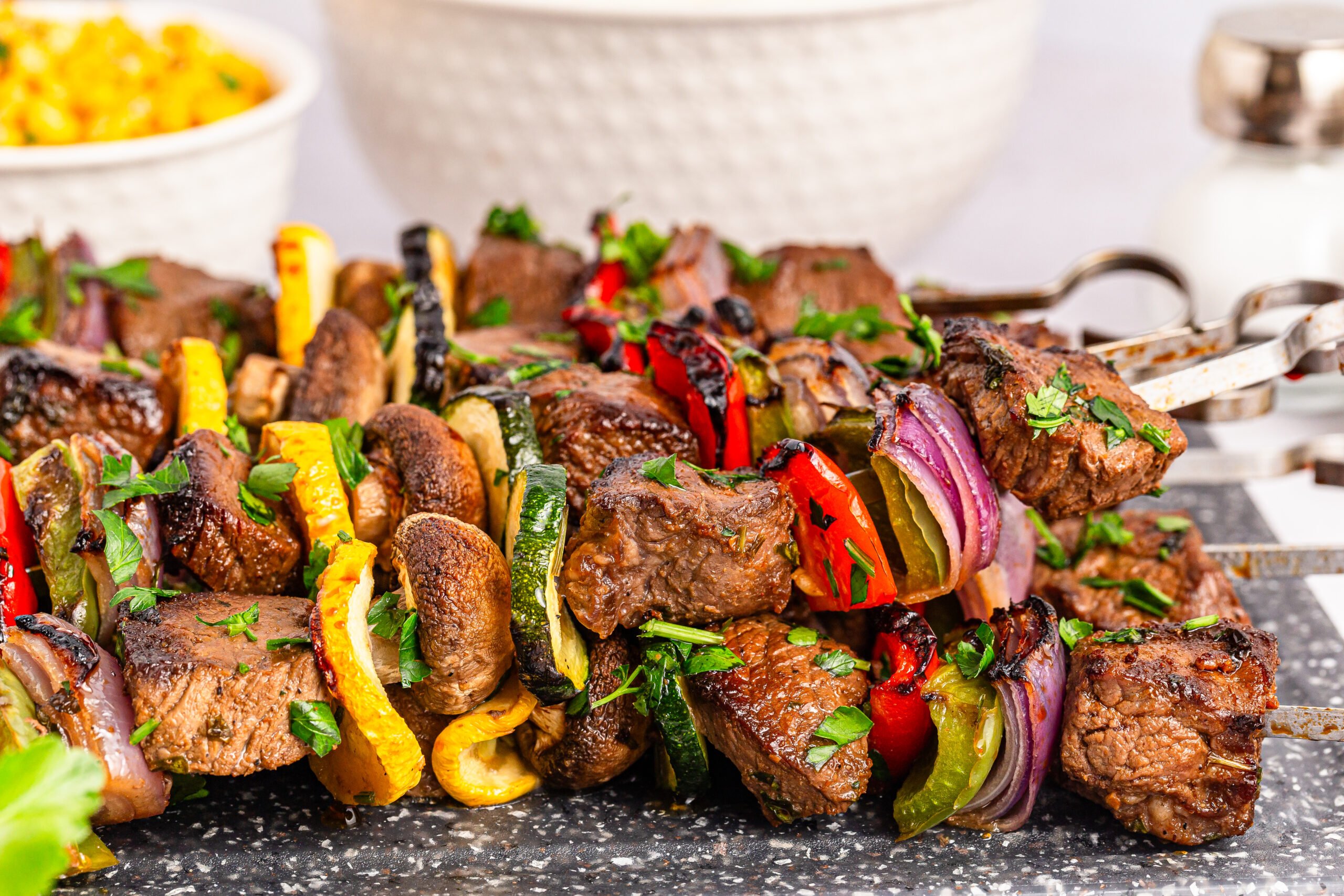 Marinated Beef Kabobs on a platter sitting on table with a bowl of corn in the background