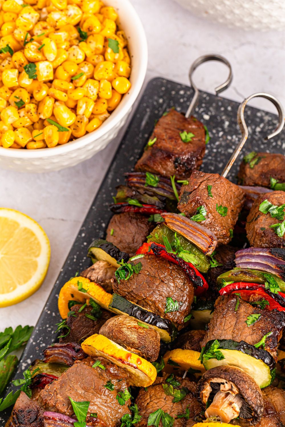 Marinated Beef Kabobs on a tray with a lemon wedge beside platter and bowl of corn 