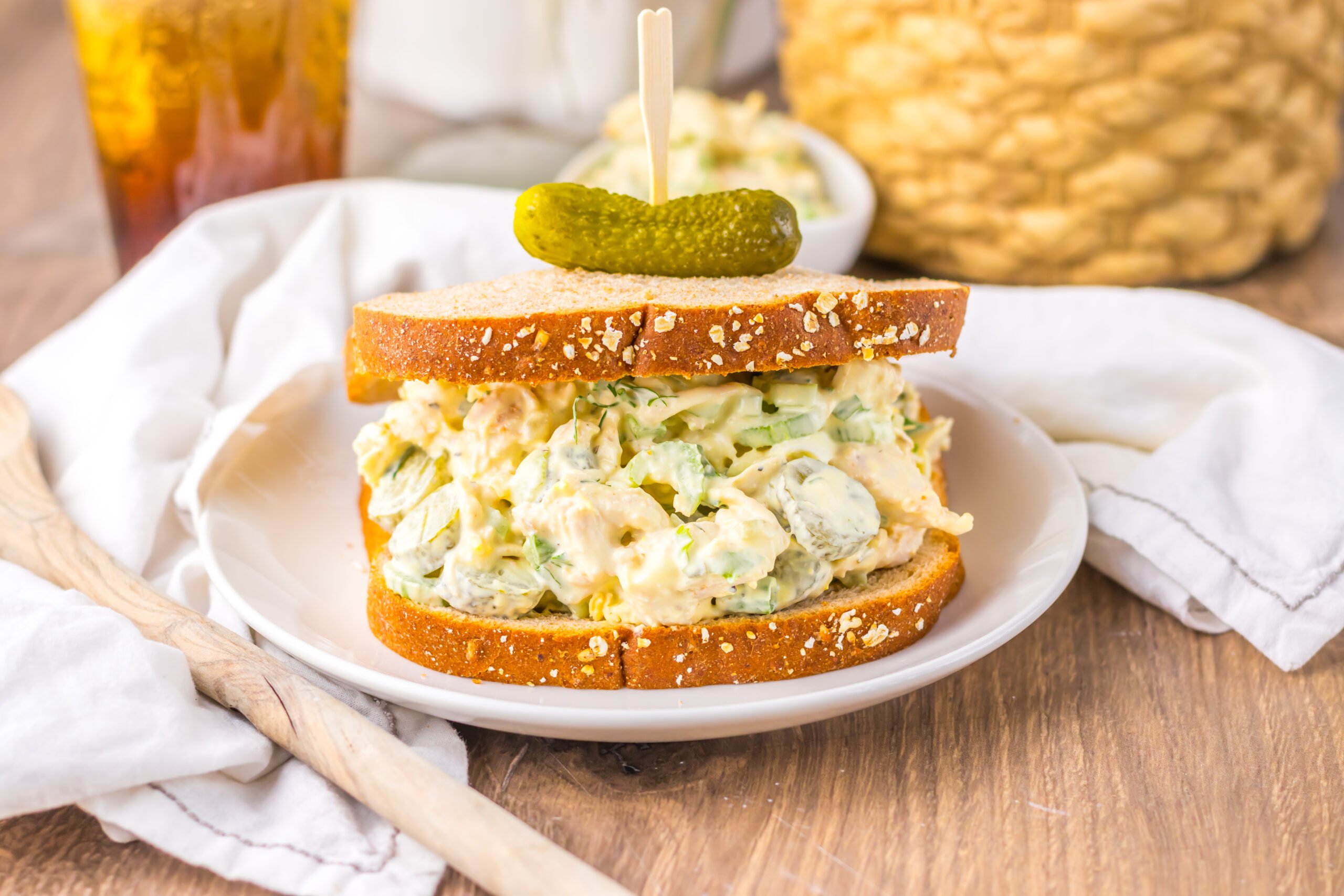 Pickle Chicken Salad sandwich on a white plate topped with a tiny pickle with a tall glass of iced tea next to it 