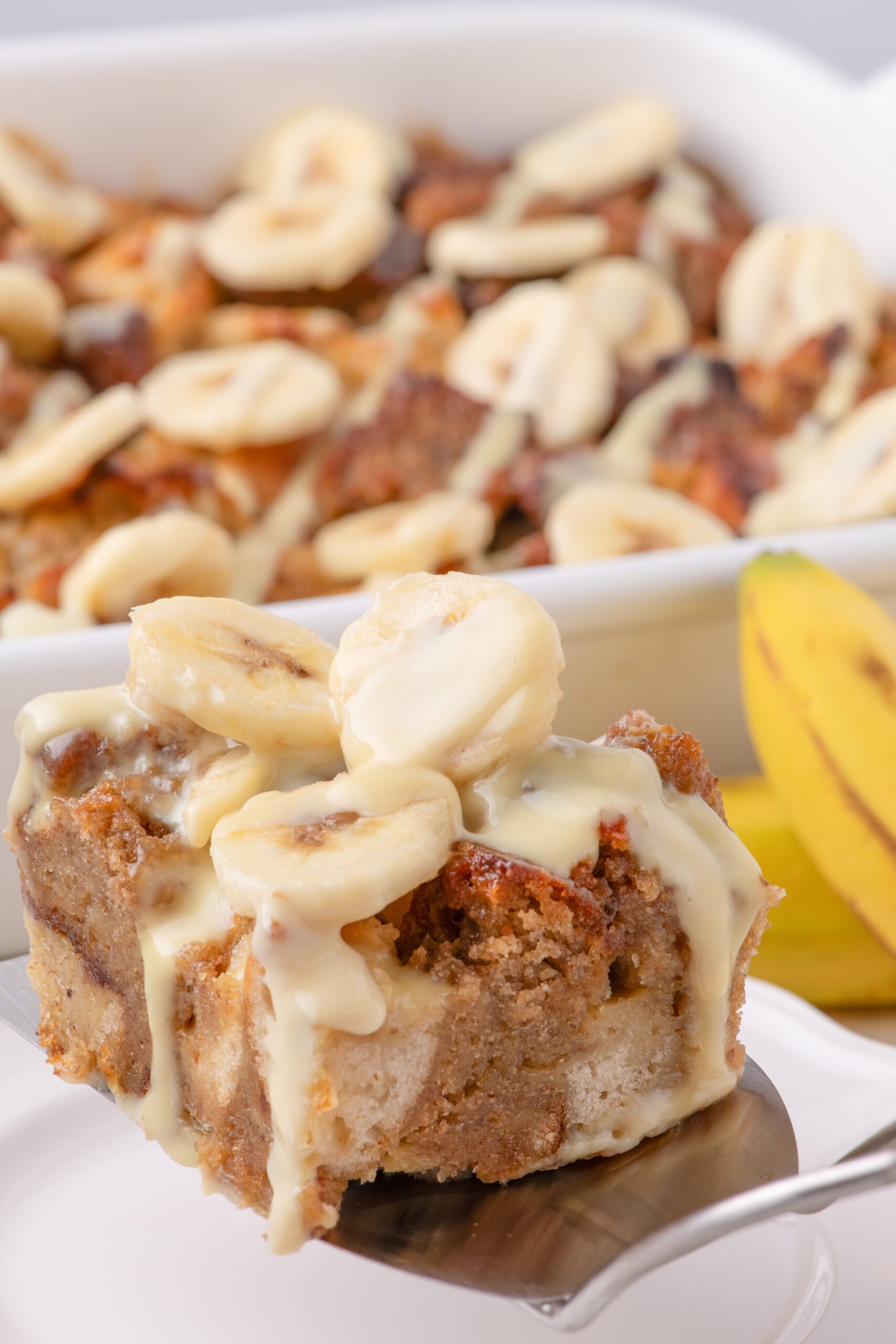 a silver spatula with a slice of Banana Bread Pudding on it topped with sliced bananas 