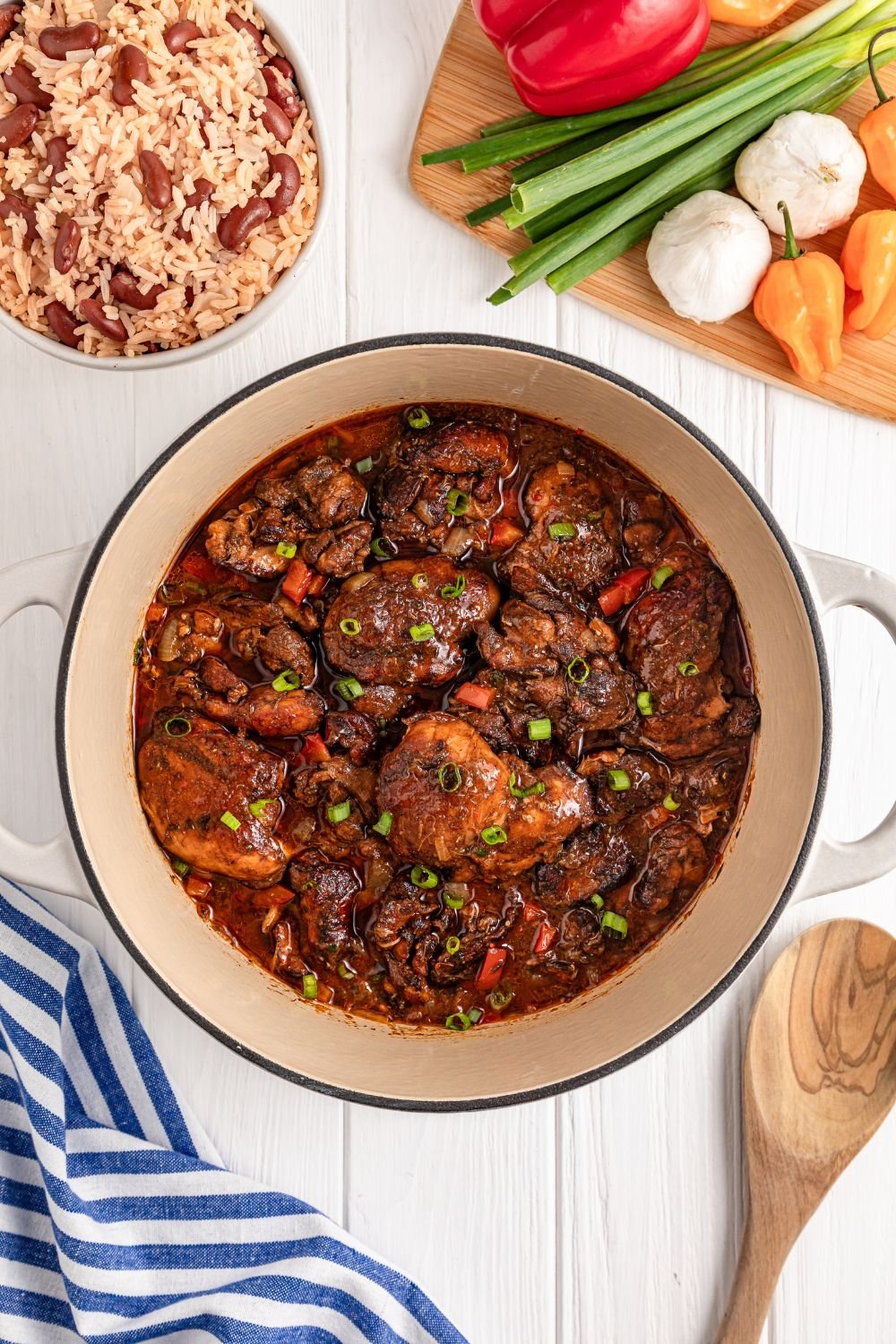 Jamaican Brown Stew Chicken in Dutch Oven sitting on table with rice and beans to side and a mix of vegetables 