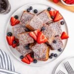 Hawaiian Roll French Toast stacked on a plate with powdered sugar and fruit on top of the French toast