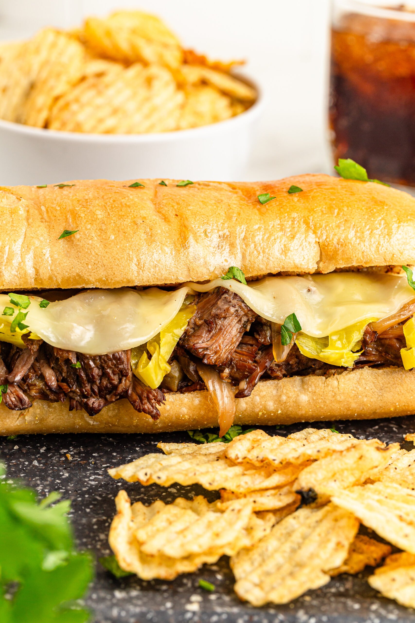 Tup close shot of a Mississippi Pot Roast Sandwich on a cutting board with a white bowl of chips and a coke in the background