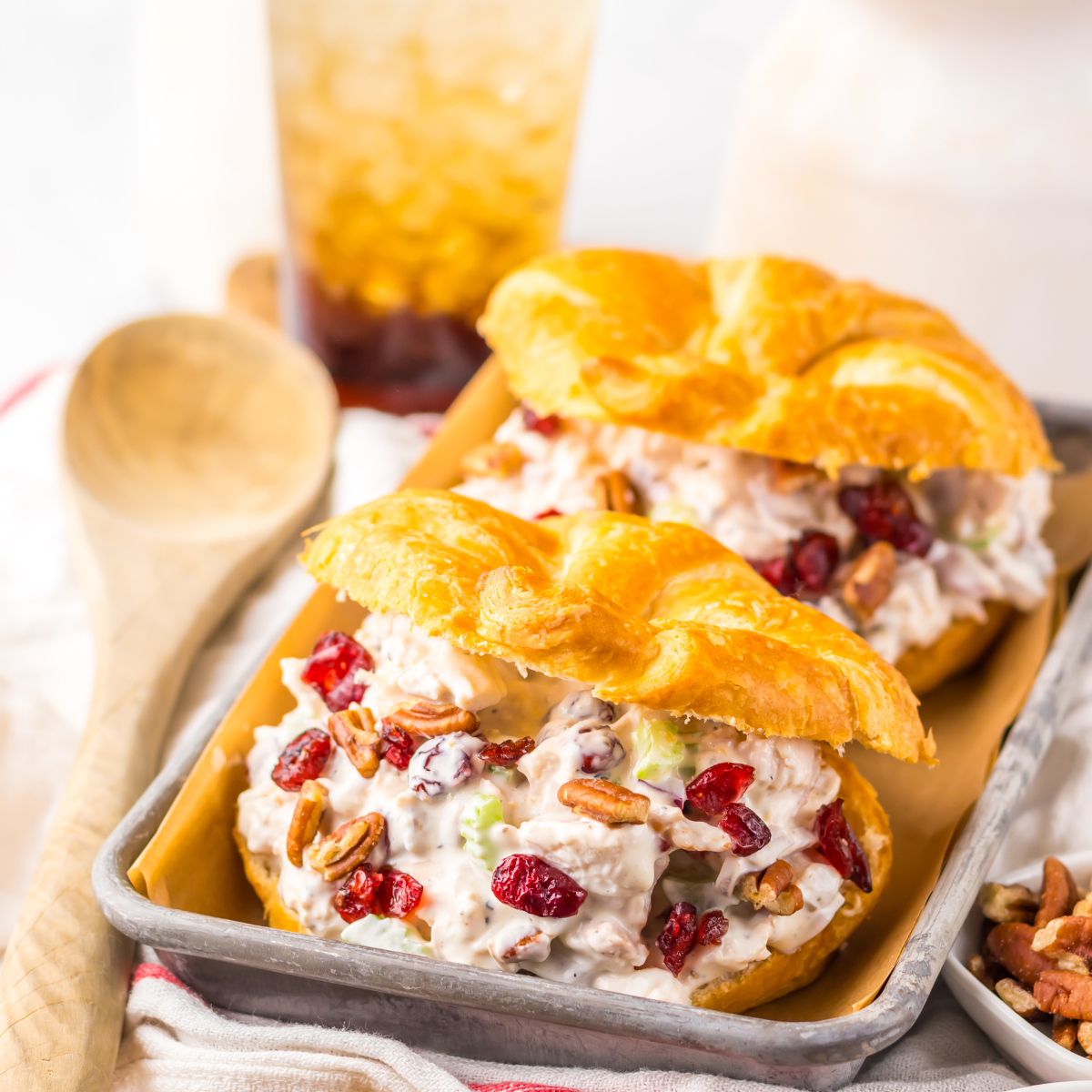 Close up view of a Cranberry Pecan Chicken Salad Croissant on a metal tray sitting on a red and white towel  with pecans and a wooden spoon tot he side.  