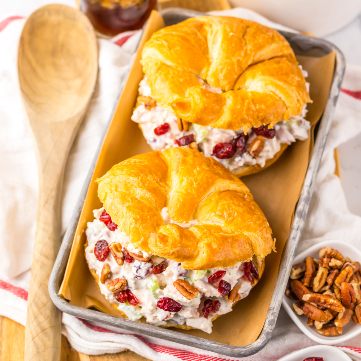 Over head view of a Cranberry Pecan Chicken Salad Croissant on a metal tray sitting on a red and white towel  with pecans and a wooden spoon to the side.  
