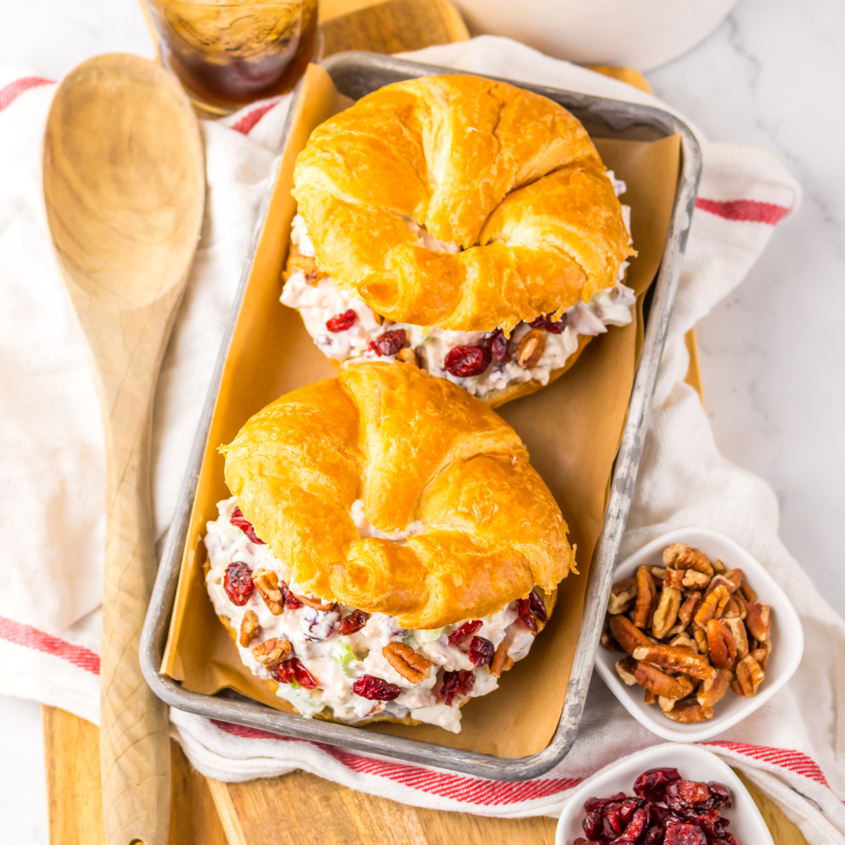 Close up view of a Cranberry Pecan Chicken Salad Croissant on a metal tray sitting on a red and white towel  with pecans and a wooden spoon to the side.  