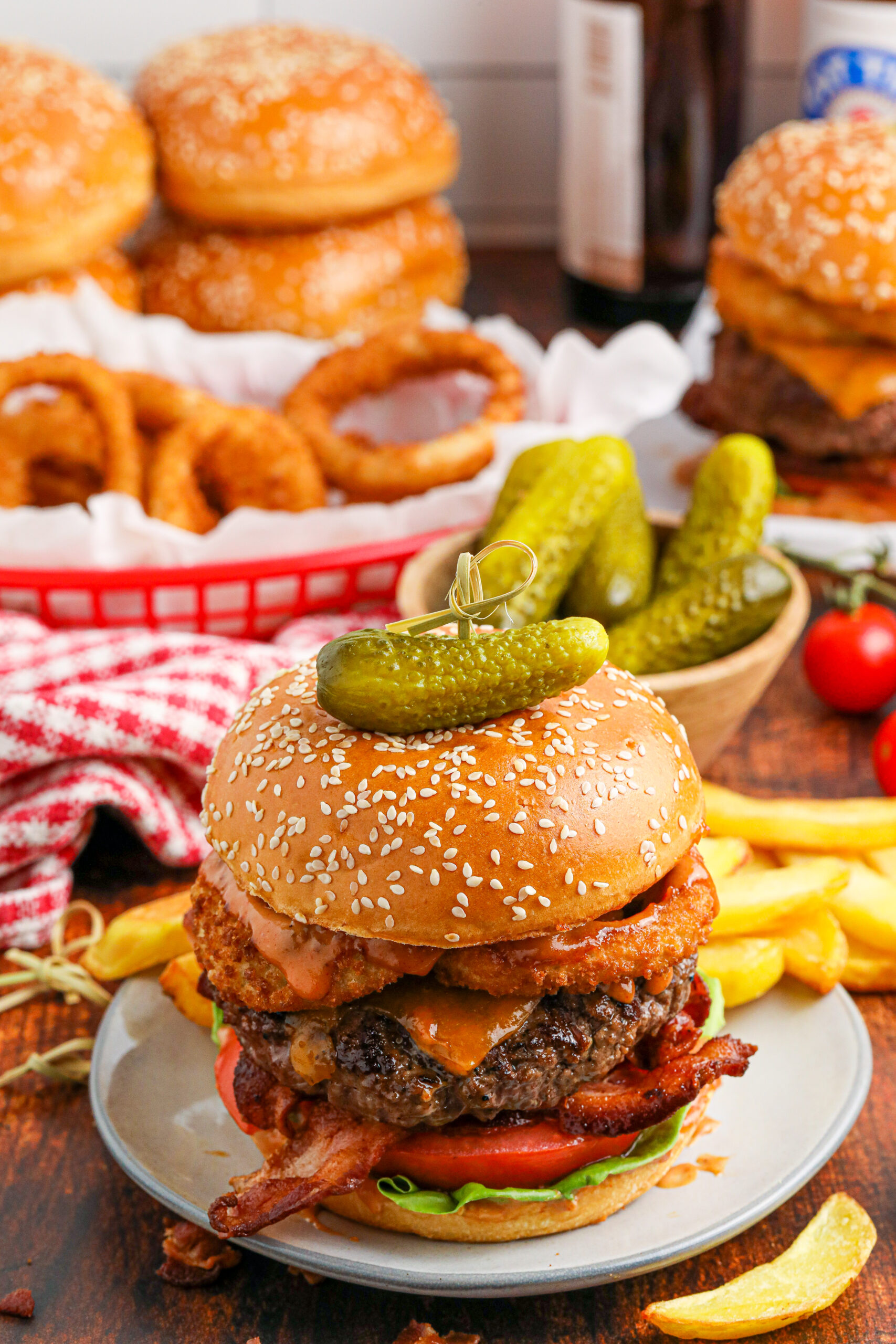 Cowboy Burger on a white plate that is fully loaded with toppings with a small bowl of pickles and fries in the background