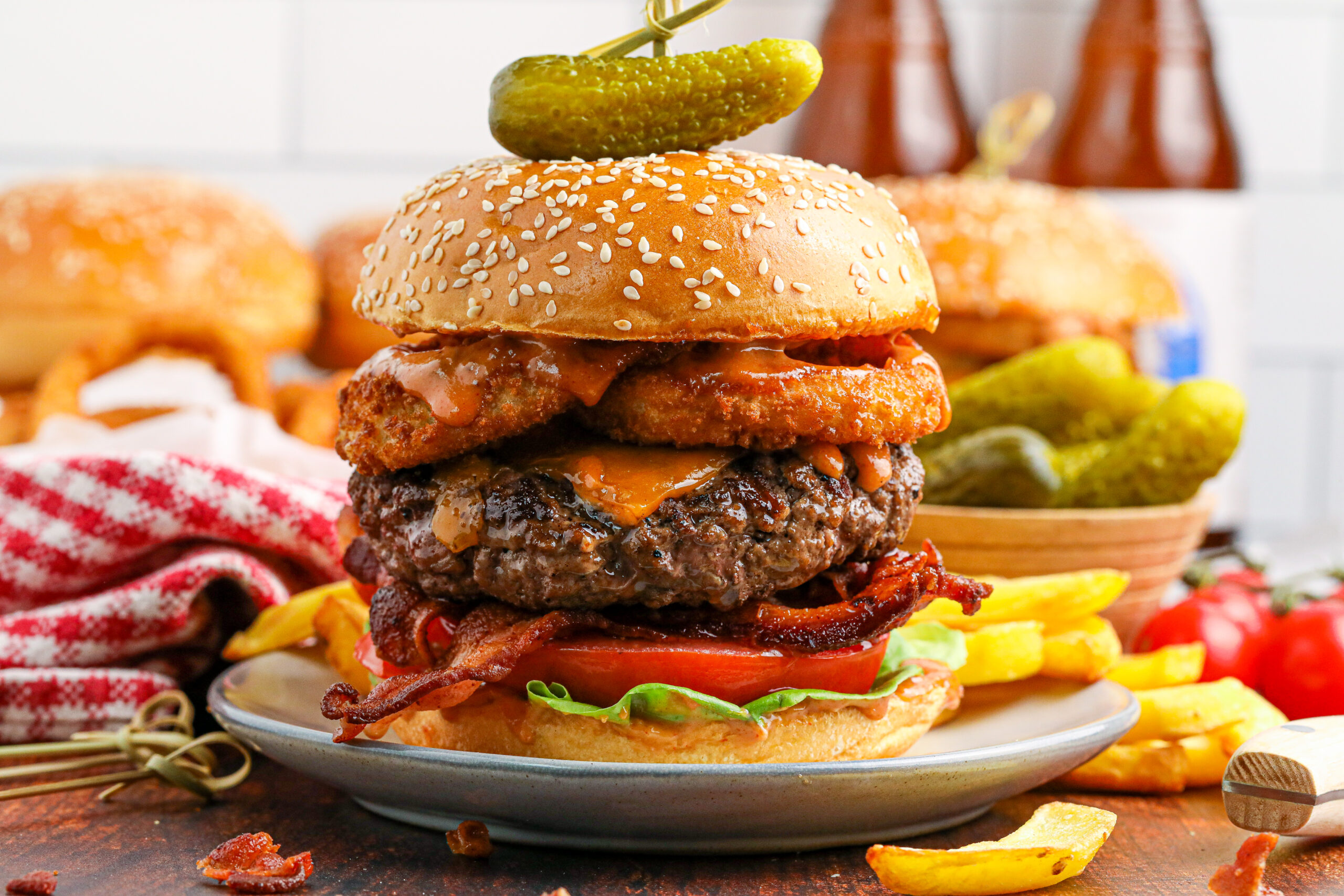 side view of the Cowboy Burger on a white plate that is fully loaded with toppings with a small bowl of pickles and fries in the background