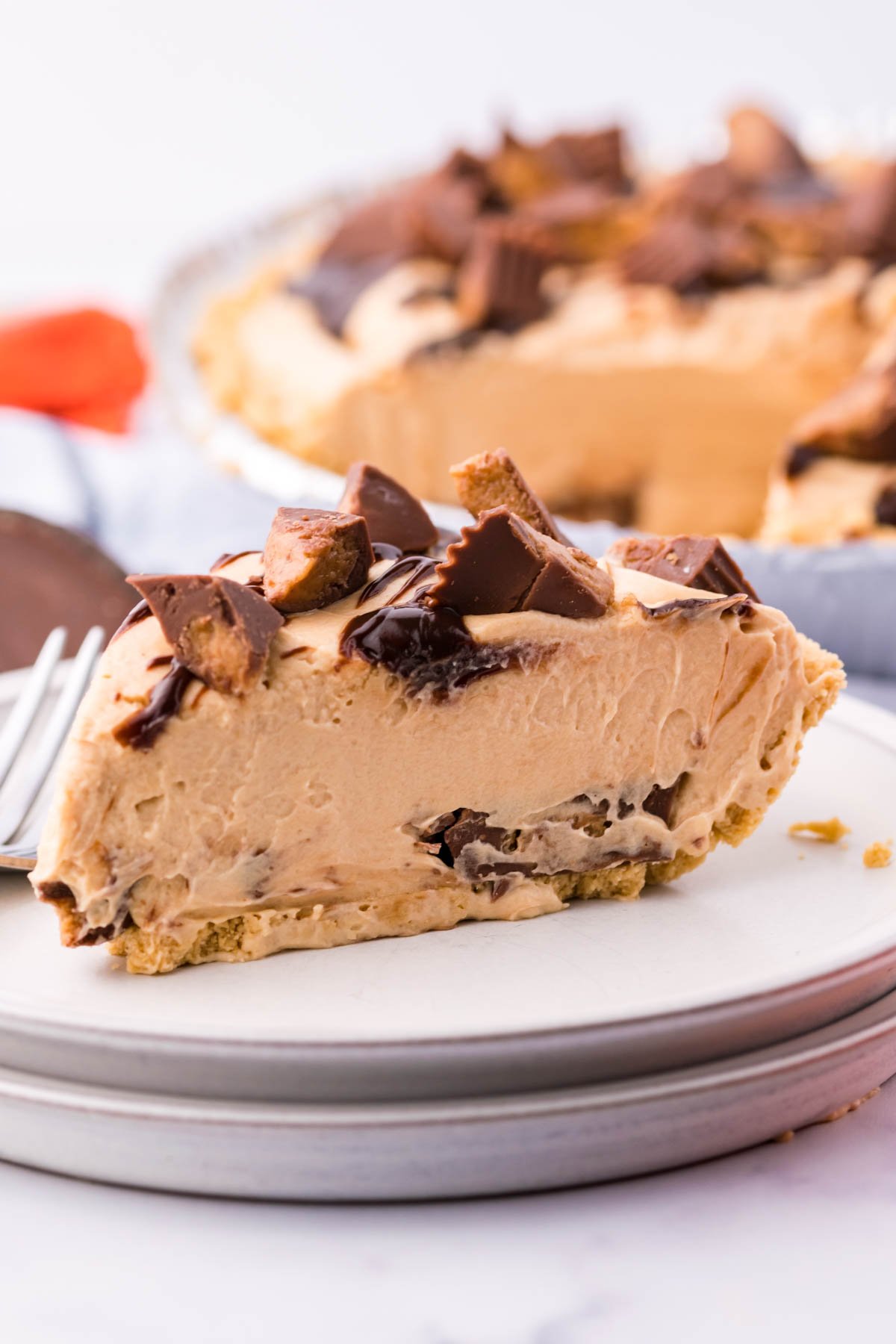 A side view of a slice of no bake peanut butter cup pie on a white plate with the full pie in the background. 