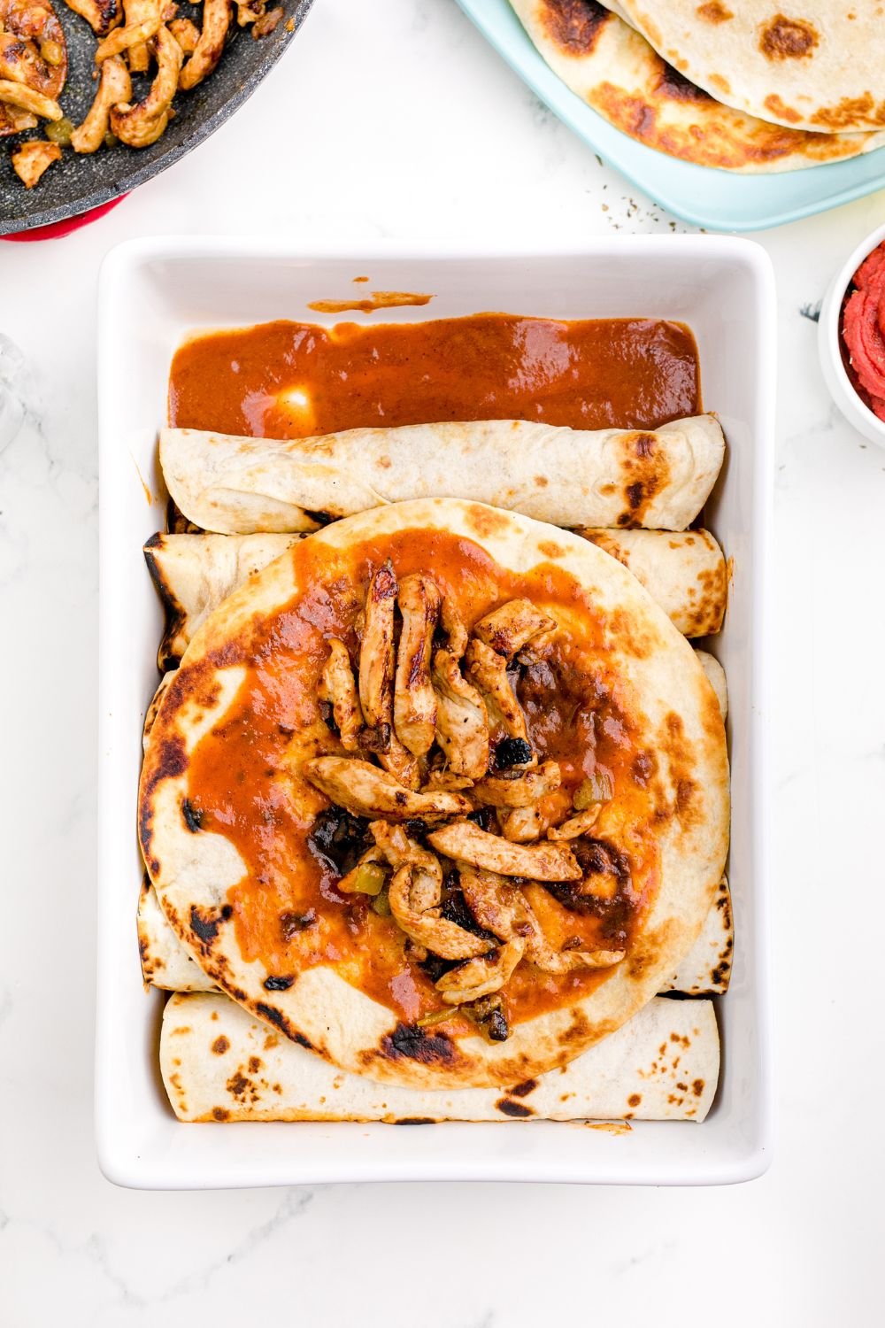 tortilla topped with enchilada sauce and cooked chicken sitting on topped of rolled enchiladas in pan 