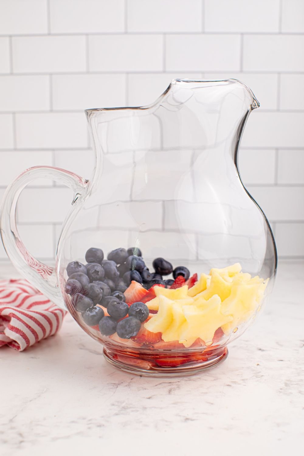 pitcher full of fruit sitting on table with a red and white towel to the side 