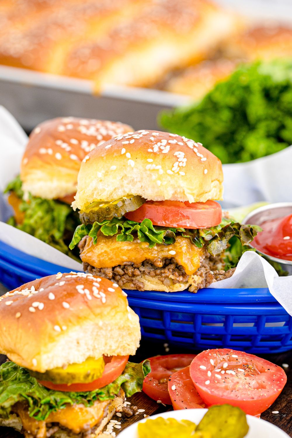 Classic Cheeseburger Sliders in baskets and spread out on table with tomatoes and ketchup around the basket, and a bowl of pickles 
