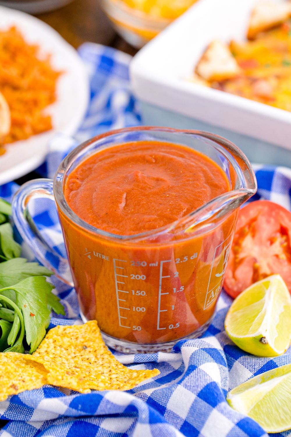 measuring cup with enchilada sauce sitting on table with tomatoes, cilantro, chips and more scattered around on table