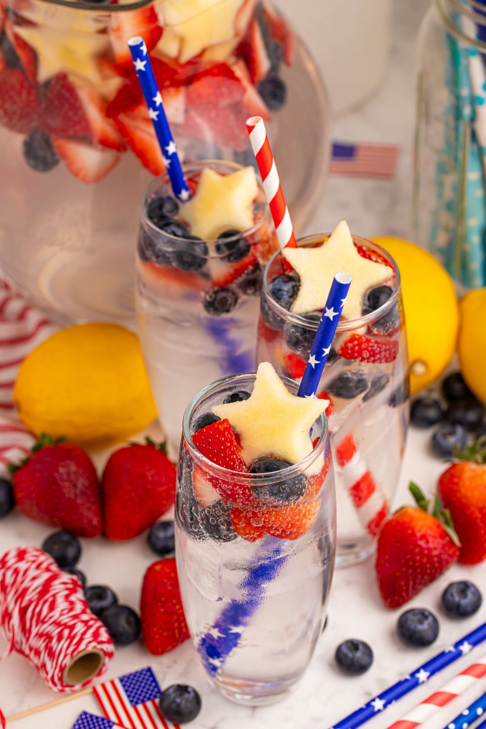 table with fresh fruit, and glasses of the Patriotic Sparkling Lemonade in it garnished with a fruit shaped like star and blue straws in each glass 