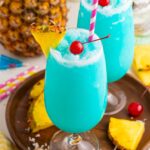 Frozen Blue Hawaiian Cocktails on serving tray with sliced pineapple and cherries around it on table