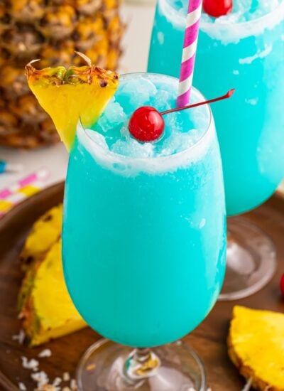 Frozen Blue Hawaiian Cocktails on serving tray with sliced pineapple and cherries around it on table
