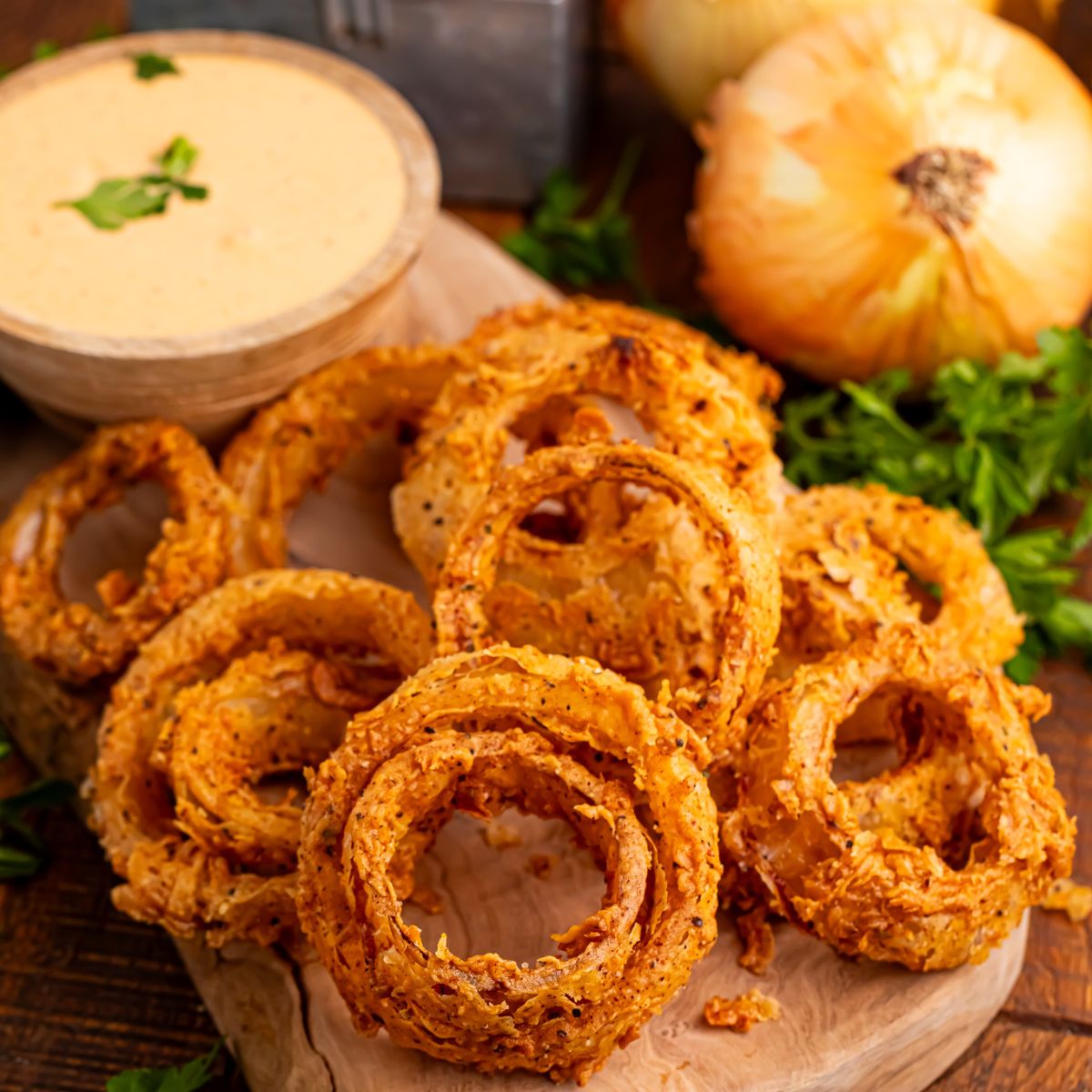 Homemade Deep Fried Onion Rings placed on a cutting board with a dipping sauce in bowl and onions on table 