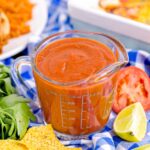 Homemade Enchilada Sauce in a measuring cup on table with tomato, lemon wedge and more around container