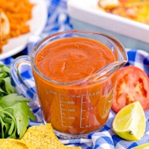 Homemade Enchilada Sauce in a measuring cup on table with tomato, lemon wedge and more around container