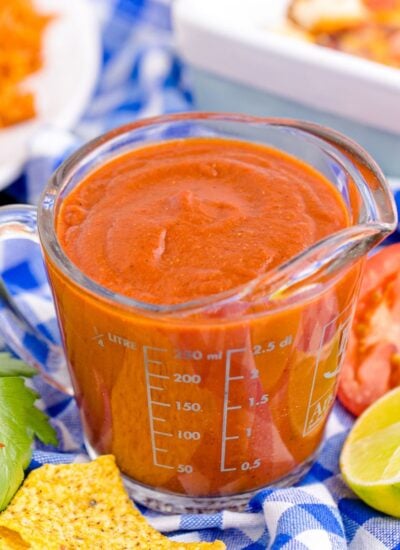 Homemade Enchilada Sauce in a measuring cup on table with tomato, lemon wedge and more around container