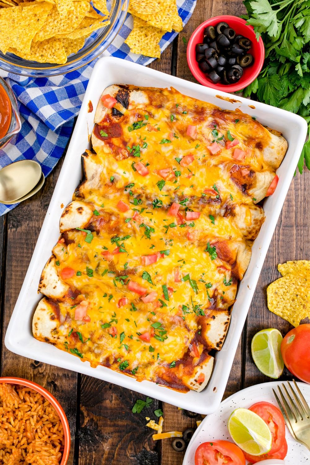overhead picture of a casserole dish full of enchiladas on table. Condiments and ingredients in bowls on table around the pan 
