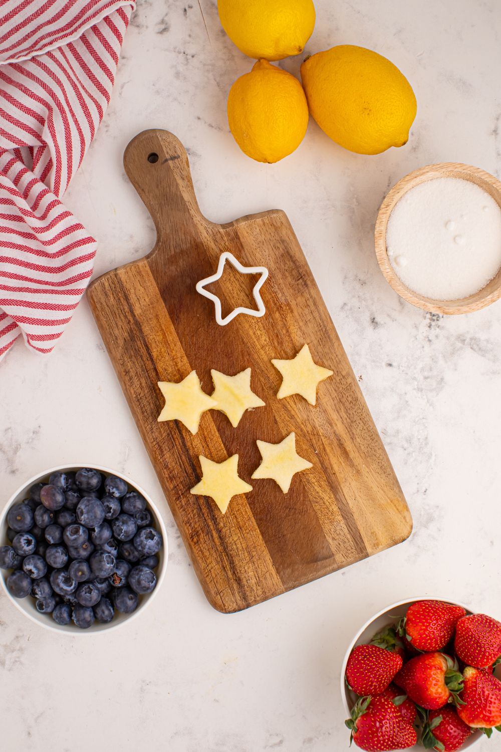 cutting board with apples cut to the shape of stars, and a bowl of sugar, blueberries ,and strawberries to the side