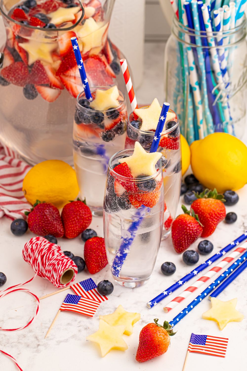 Patriotic Sparkling Lemonade in glasses with pitcher of lemonade to side. Fresh fruit scattered around on table with little American flags, and ribbon